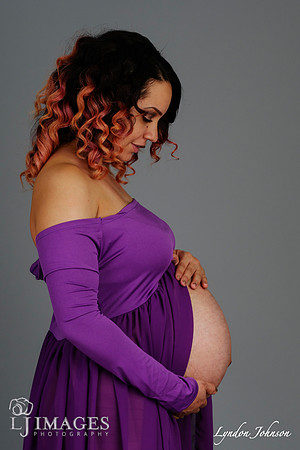 studio maternity portrait in purple dress, belly exposed, looking down at belly, on grey background