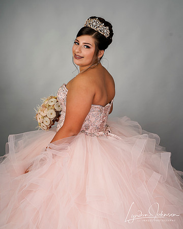 studio quinceañera portrait on grey background wearing pink dress and holding pink roses, looking back over her shoulder